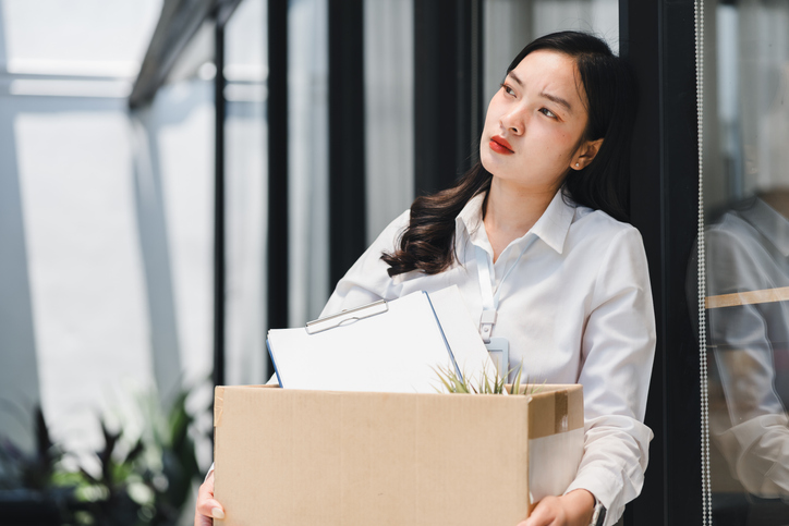 A disheartened young business woman carrying a box of her belongings, feeling sad about being fired or resigning from her job. hive-blockchain-technologies-layoffs-know-your-rights